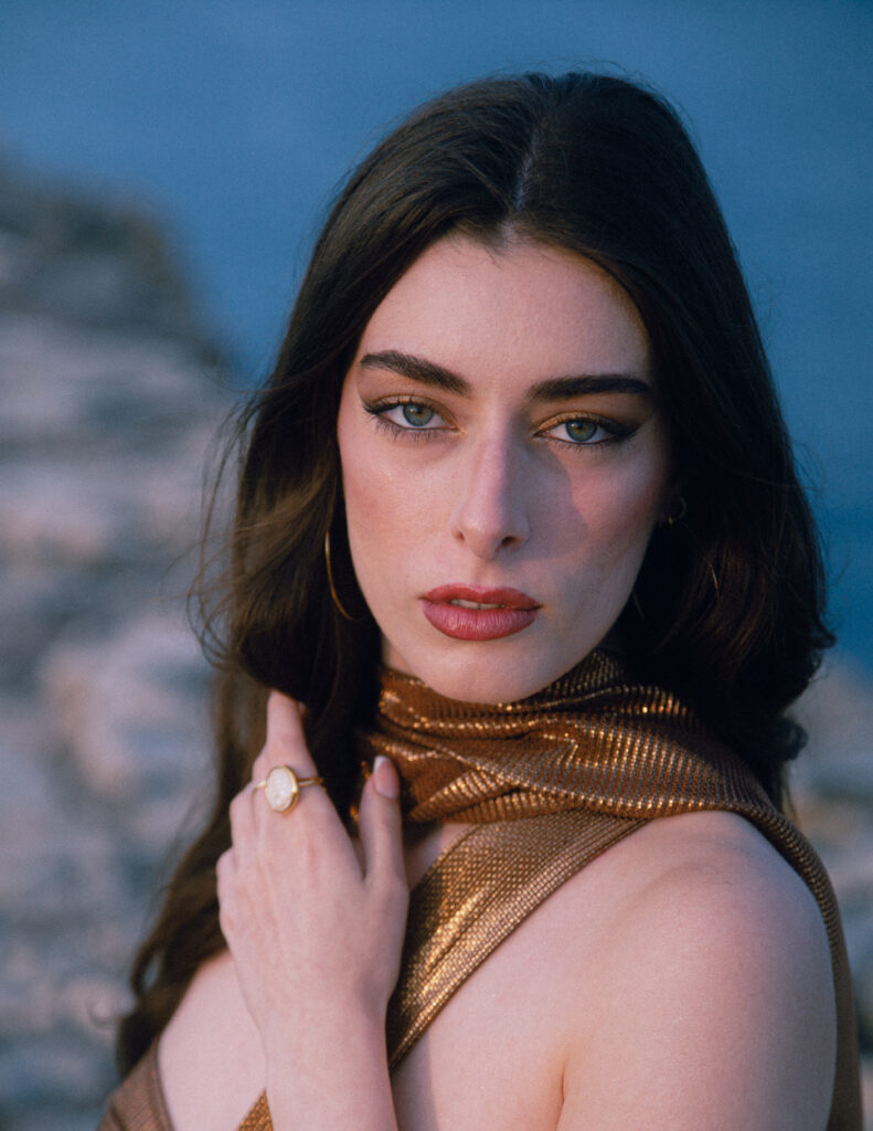 Close up portrait photograph of a beautiful young woman. Big eyes and lips, a golden ring and earrings, she's wearing a gold colored textured top and scarf. All around her is the blue sea.