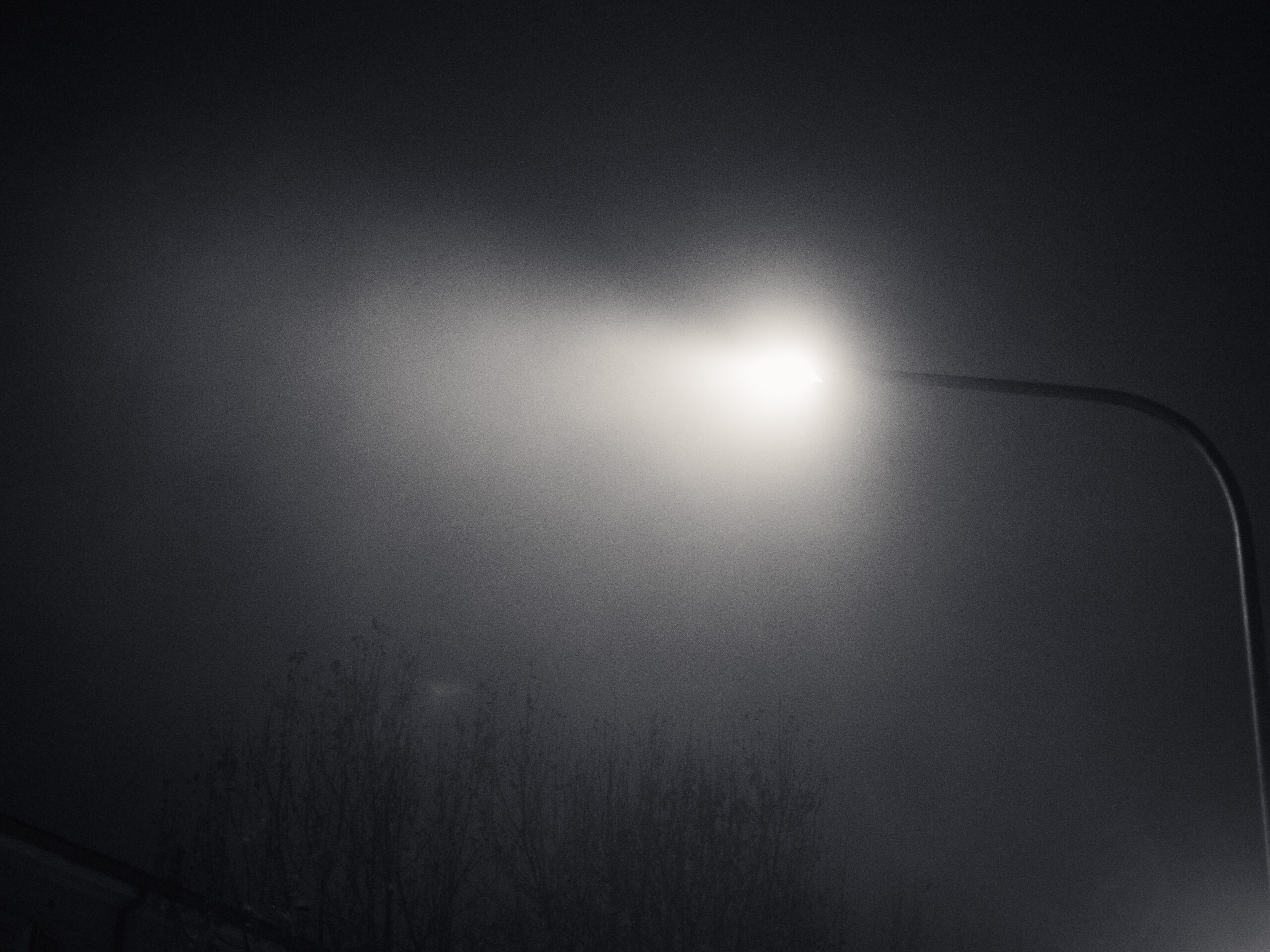 Grainy black and white photograph of a streetlamp, the light diffused by humidity.