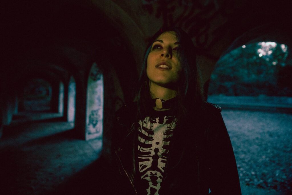 Dark close photograph of a punk girl with blue hair leaning against the wall of a long empty archway.