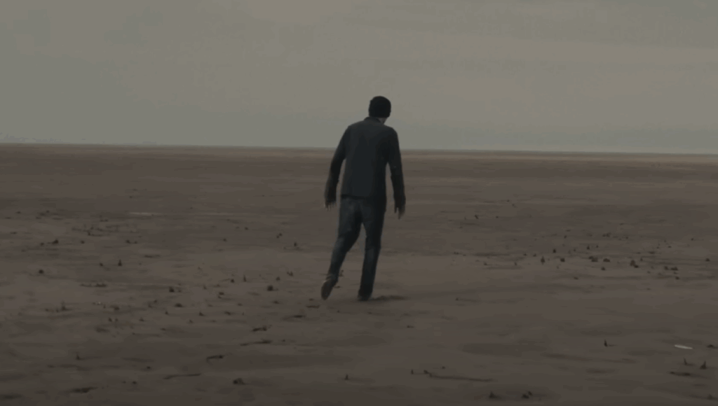 Still from Sleep Token's 'Fall For Me' music video. The protagonist is wandering in an empty expanse of wet sand.